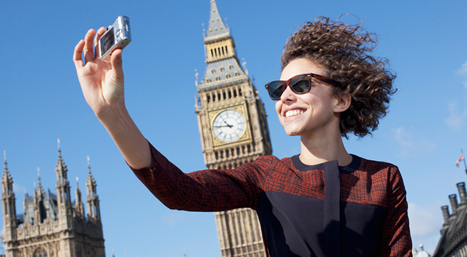 Woman with curly hair in Wayfarer sunglasses taking photo with digital camera in front of Big Ben
