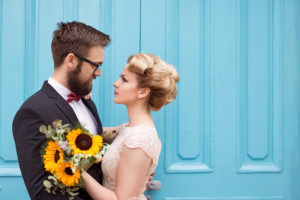 Beautiful vintage bridal wedding style Newlywed couple standing next to a blue retro wooden door, holding a sunflower wedding bouquet and looking at each other